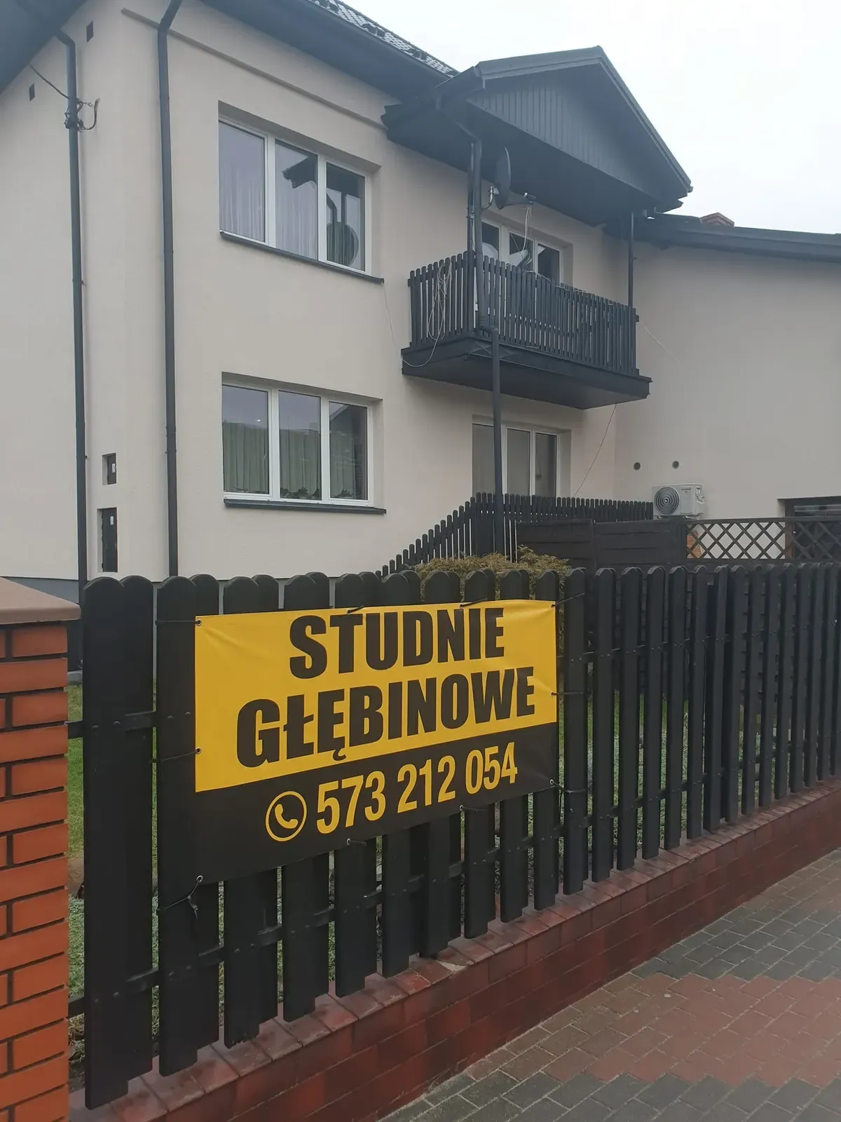 Żółto-czarny baner firmy Studnie Głębinowe z numerem telefonu 573 212 054 na ogrodzeniu domu
